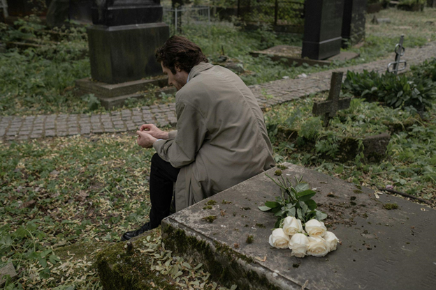 A person sits solemnly on a grave, hands clasped, beside white roses.